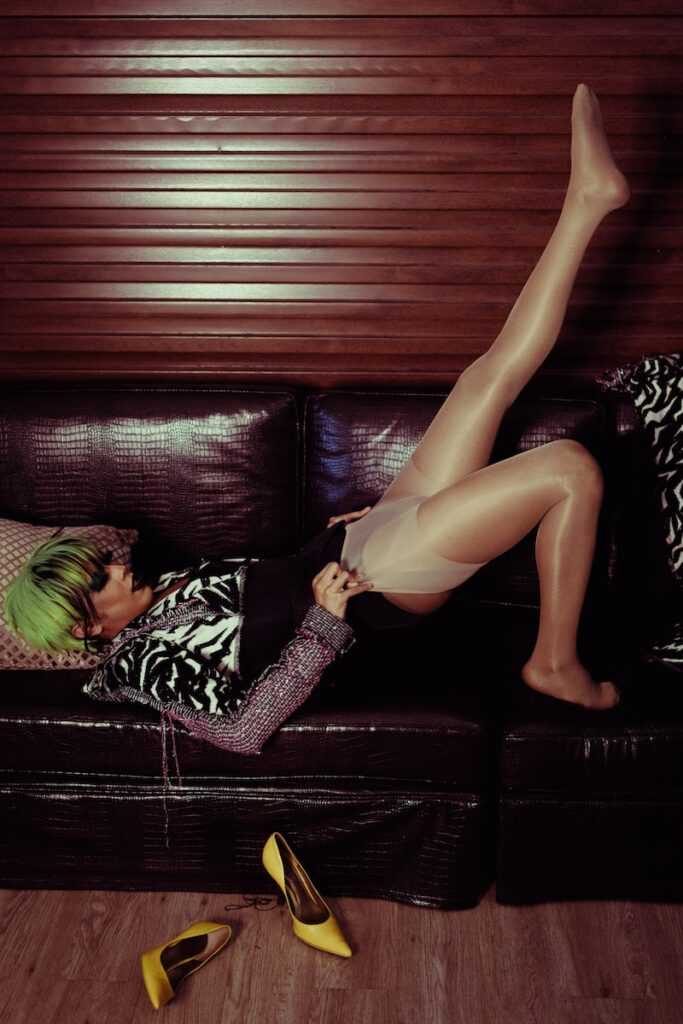Side view of young stylish female with bright makeup putting on nylon tights while lying with raised leg on sofa